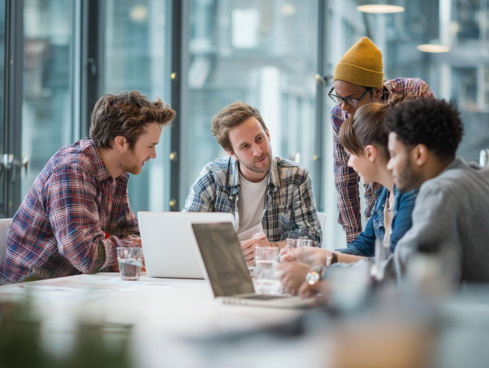 A diverse group of young professionals collaborating around a table with laptops and glasses of water in a modern office setting.